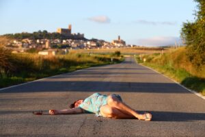 A woman in a light blue dress is lying motionless on a deserted road, with a pair of sunglasses nearby. The background shows a distant village with historic buildings, surrounded by fields and greenery. The scene suggests a hit-and-run accident.