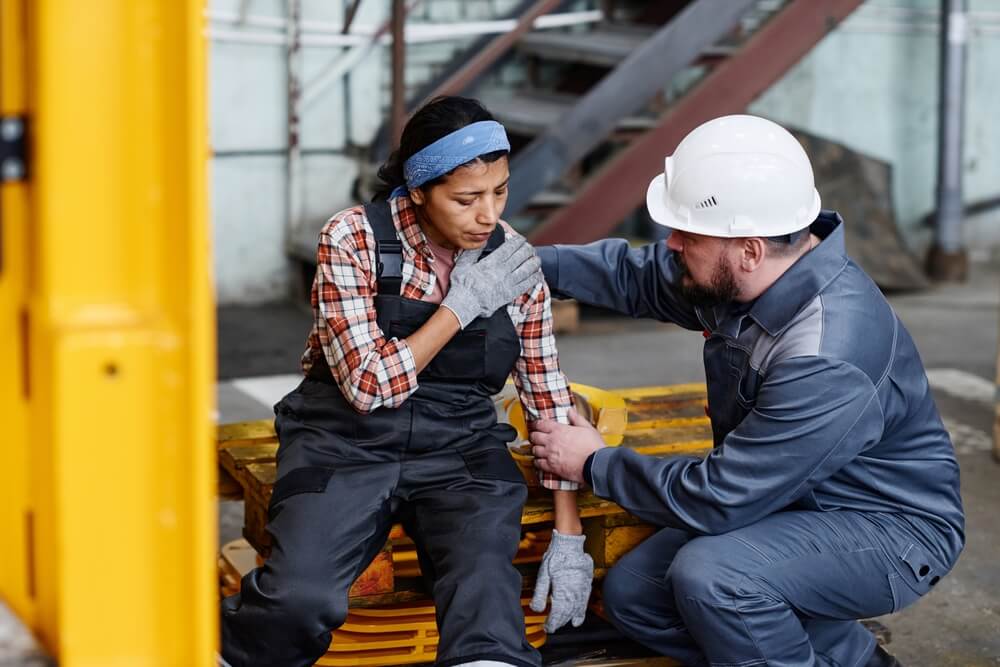 Mature foreman in workwear giving first aid to female subordinate with pain in her shoulder after accident at work in warehouse